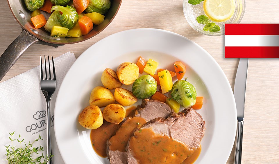 Ein Teller mit einem klassischen Schweinsbraten mit Bratkartoffeln und Gemüse auf einem gedecktem Tisch. Ein Teller mit einem klassischen Schweinsbraten mit Bratkartoffeln und Gemüse auf einem gedecktem Tisch.